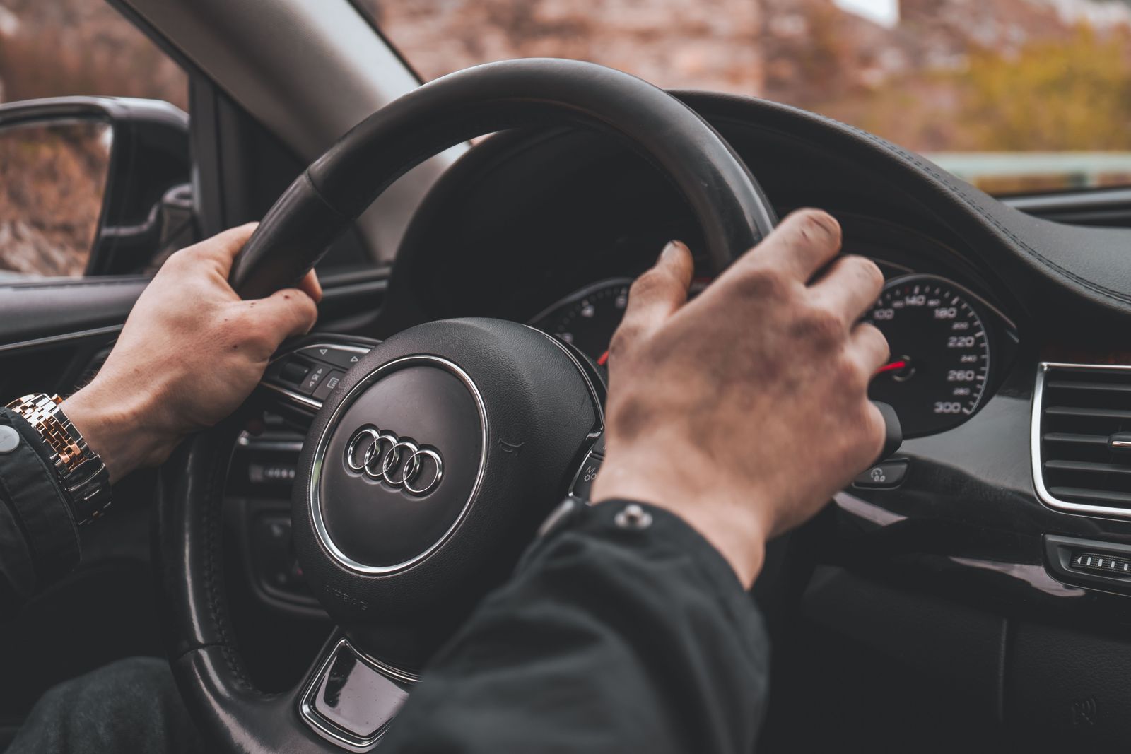 A driver's hands on the steering wheel of a premium car, with a dark suit sleeve and a wristwatch