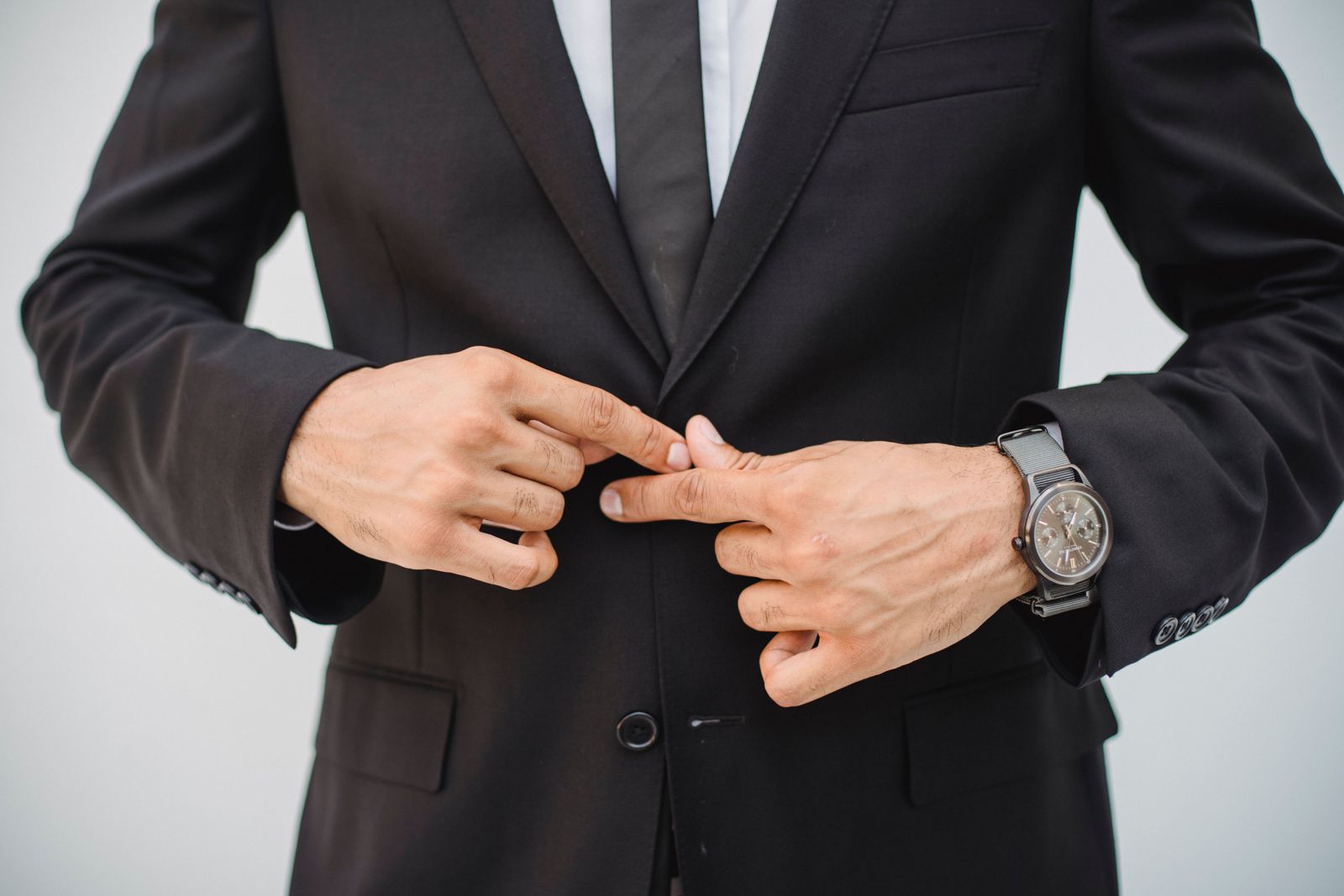 Close-up of a man's hands buttoning a dark suit jacket, wristwatch visible